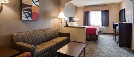 A hotel room with a sofa, a small table, a desk with a computer, a TV, and a bed with a red blanket.