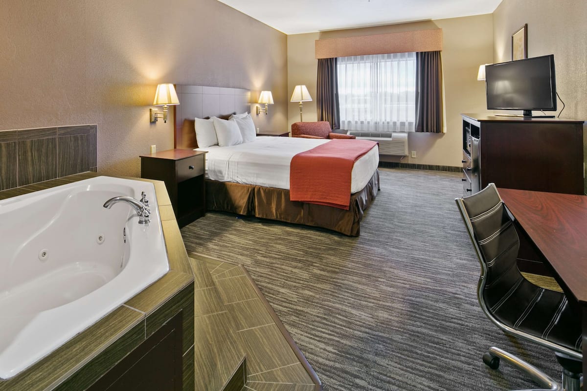 A hotel room with a large bathtub, a bed with a red blanket, a desk with a TV, and a window with curtains.