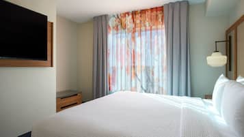 A hotel room with a large bed, a flat-screen TV mounted on the wall, a bedside table, and a window with sheer curtains.