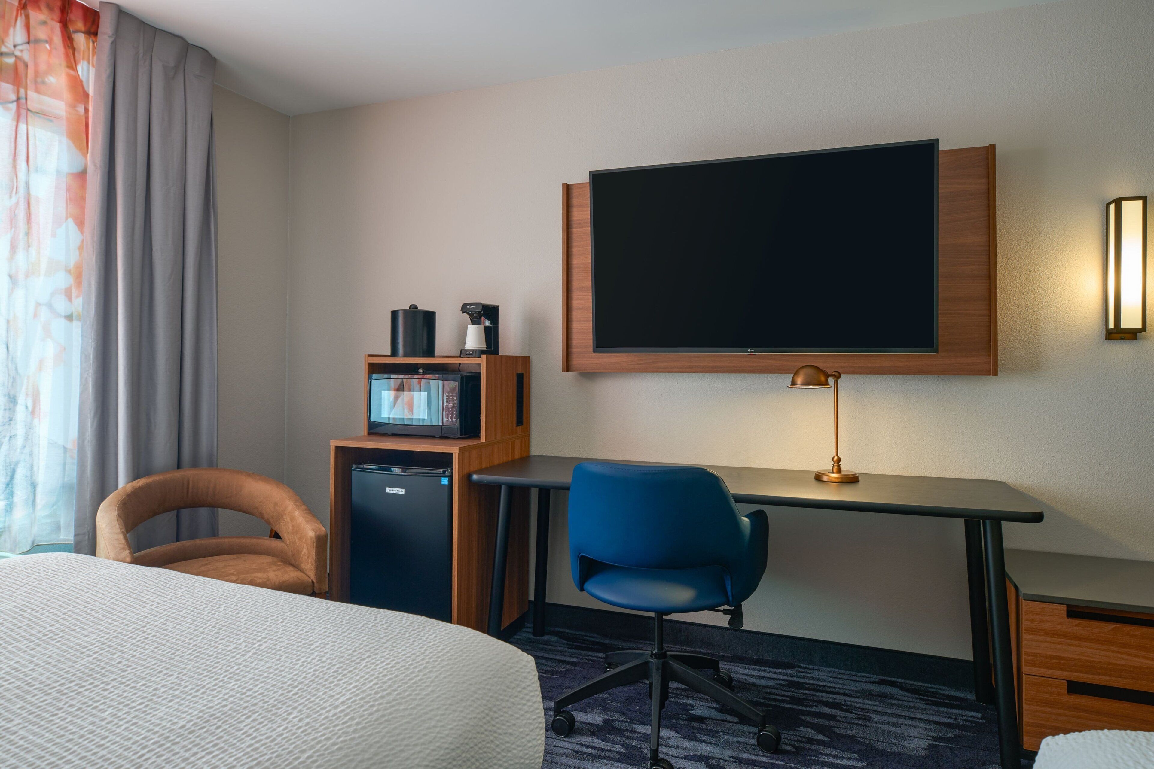 Una habitación de hotel con una cama, un escritorio con una computadora, una silla, un televisor montado en la pared y un refrigerador pequeño.