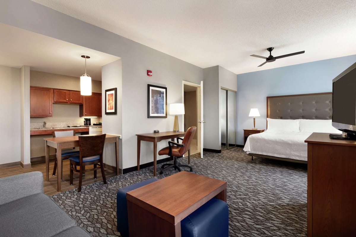 A hotel room with a bed, desk, chair, kitchenette, and a ceiling fan.