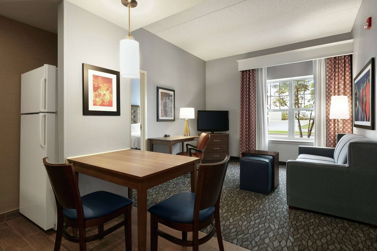 A hotel room with a dining table, two chairs, a sofa, a TV, and a window with curtains.