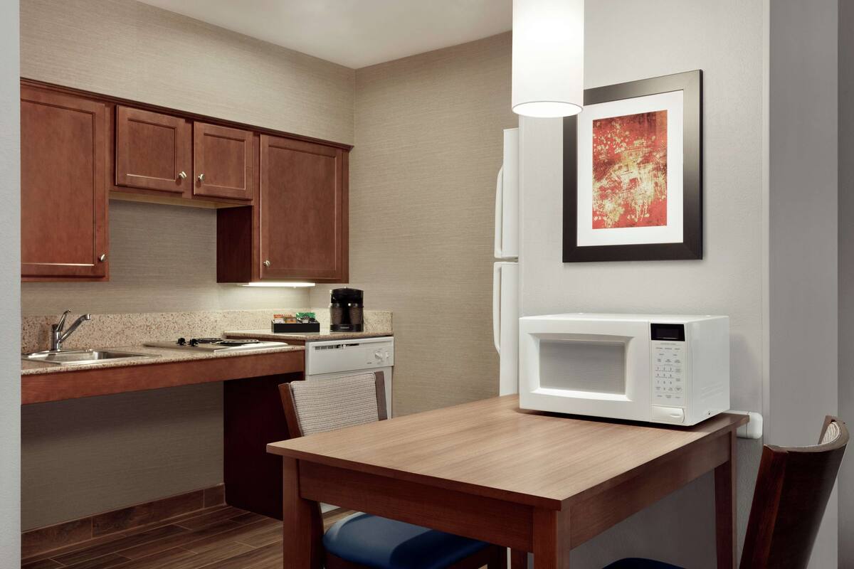 A compact kitchen with wooden cabinets, a microwave, a coffee maker, and a dining table with a chair.