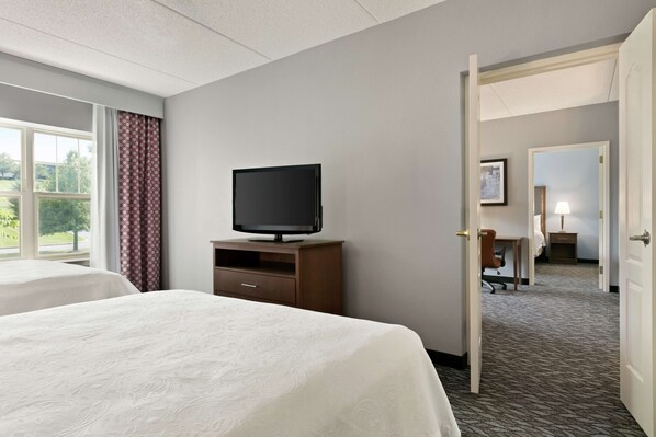 A hotel room with a bed, a television on a wooden stand, a desk with a chair, and a door leading to another room.