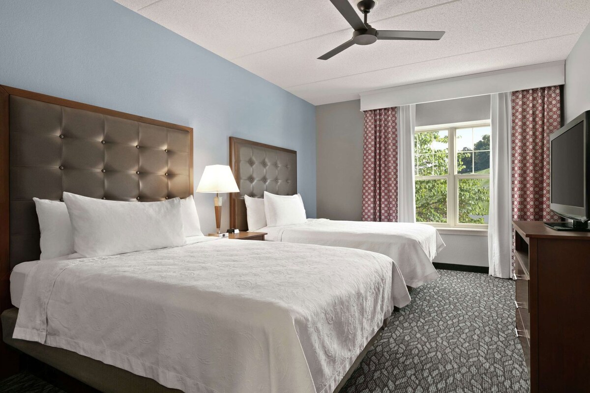 A hotel room with two beds, a ceiling fan, a TV, and a window with curtains.