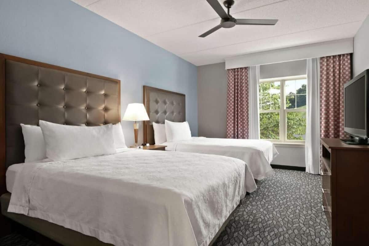 A hotel room with two beds, a ceiling fan, a TV, and a window with curtains.