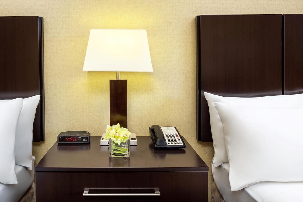 A hotel room with a nightstand, a digital clock, a lamp, a vase with flowers, a telephone, and two white pillows.