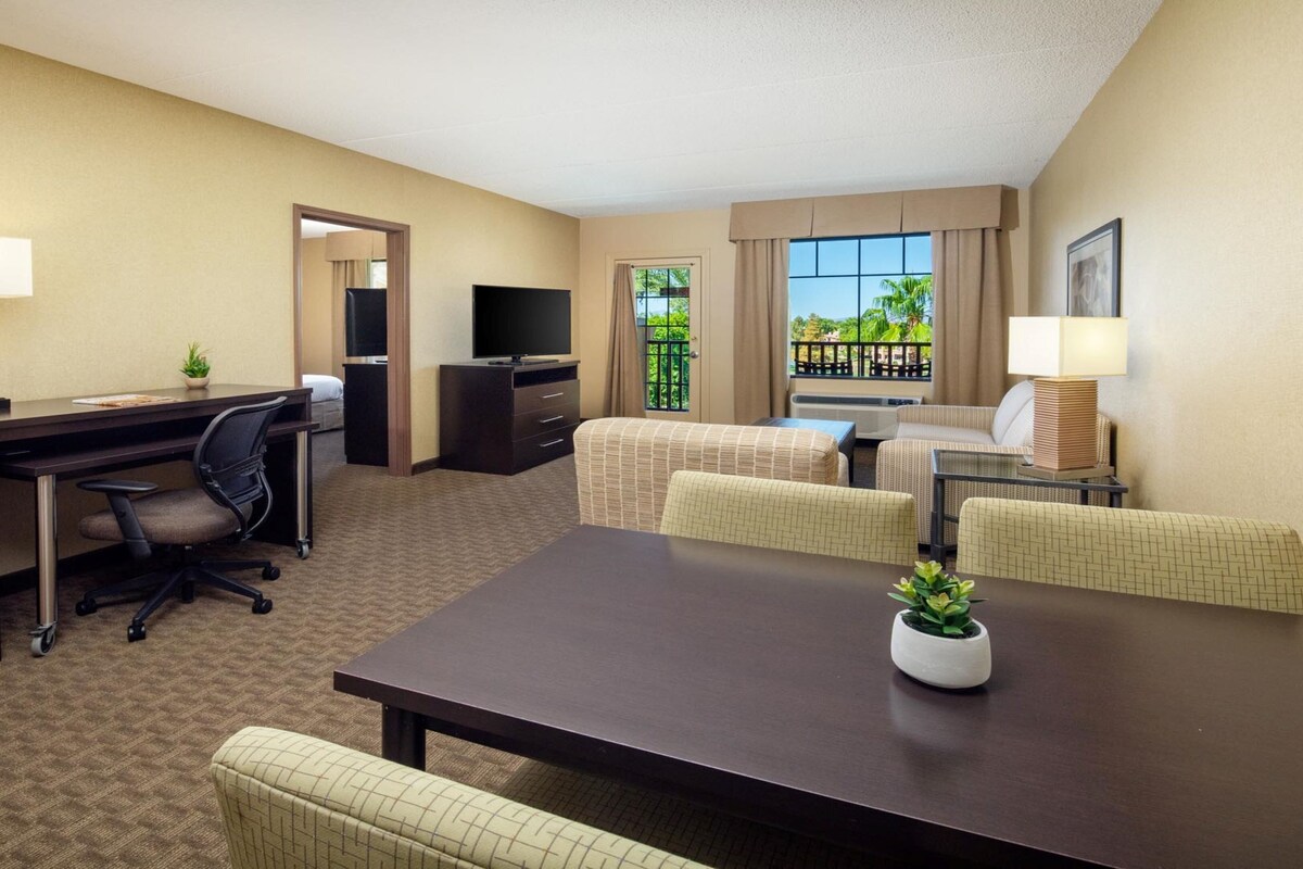 A hotel room with a desk, office chair, two armchairs, a sofa, a TV, and a window with a view of greenery.