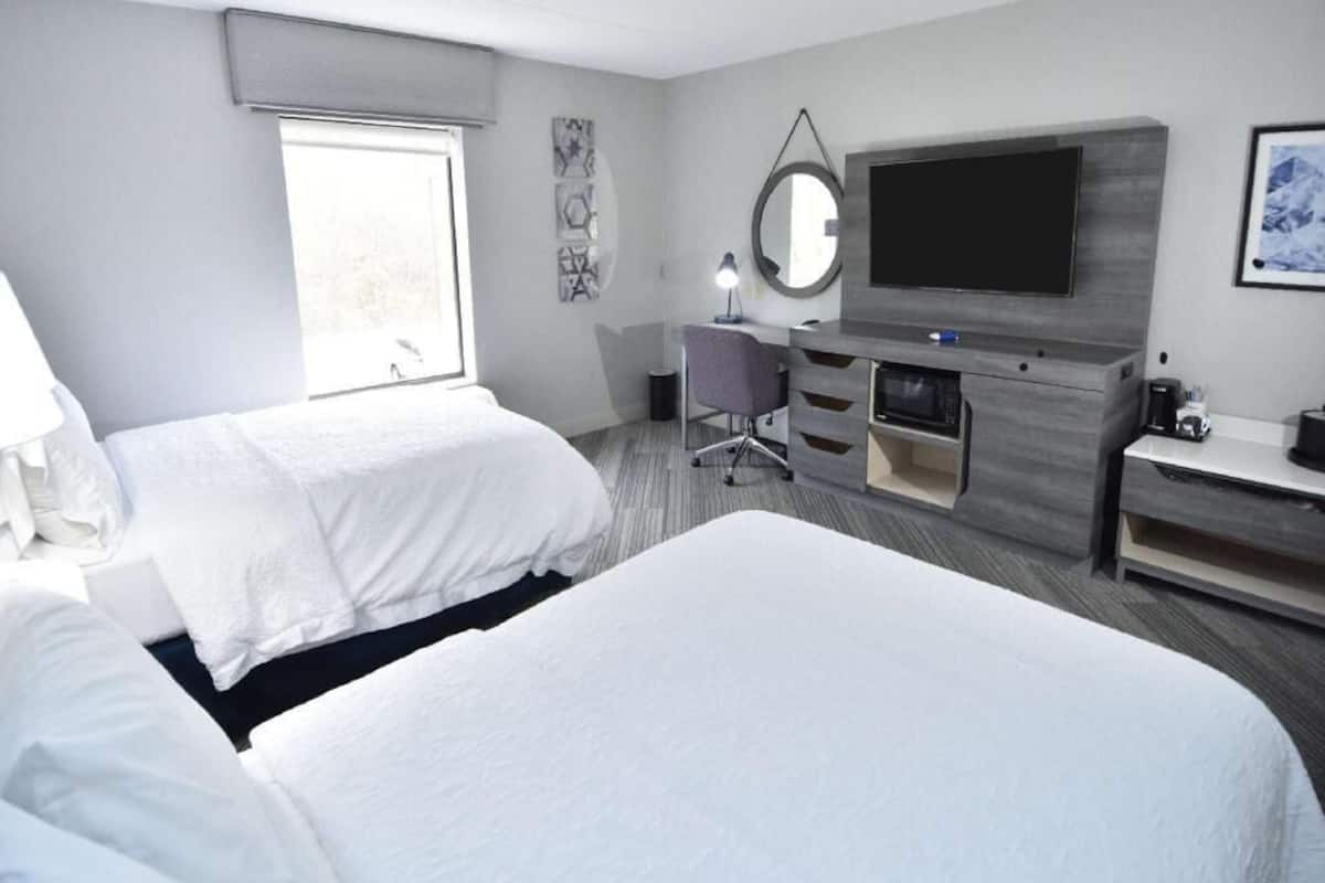 A hotel room with two beds, a desk, a television, and a window.