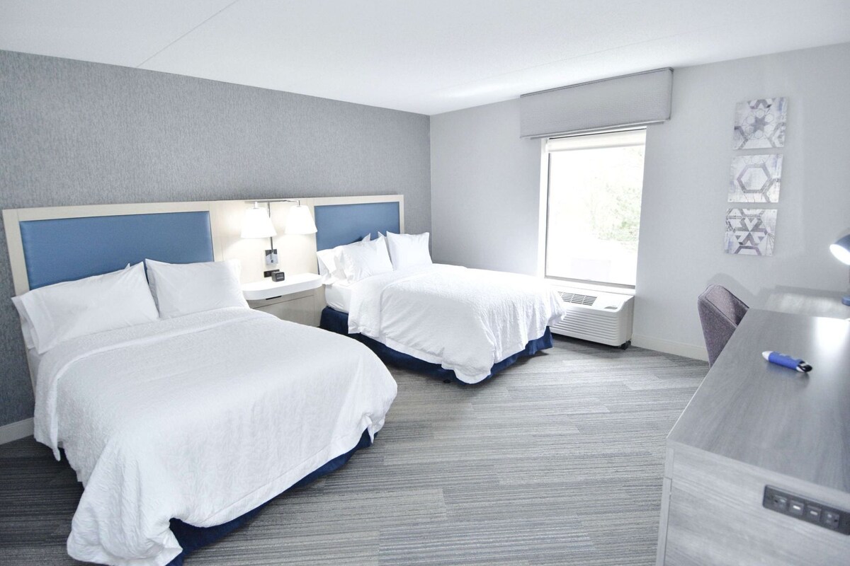 Habitación de hotel con dos camas, un escritorio, una ventana y un aire acondicionado de pared.