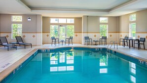 A swimming pool with lounge chairs and tables, surrounded by windows with a view of greenery.