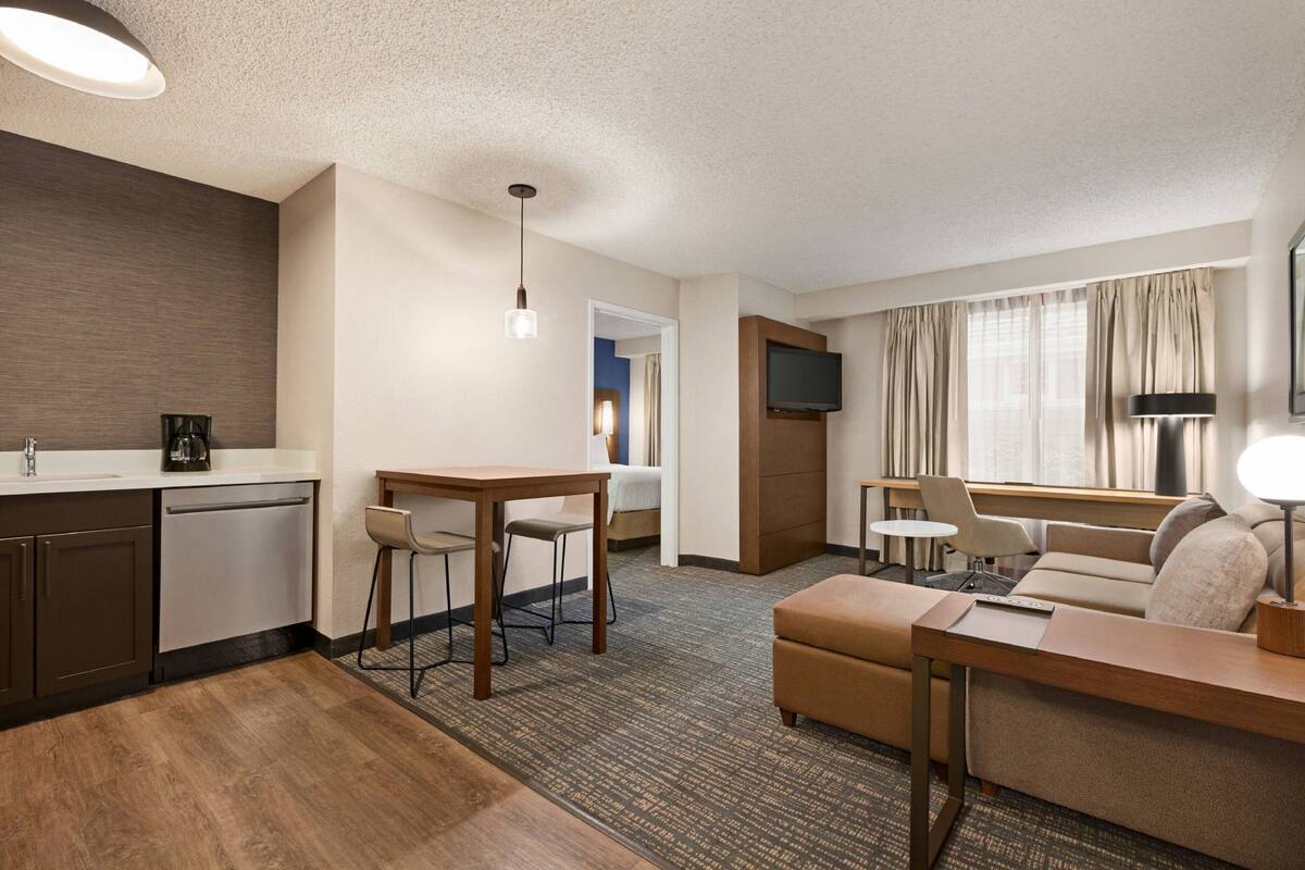 A hotel room with a kitchenette, a dining area, a sofa, a TV, and a window with curtains.
