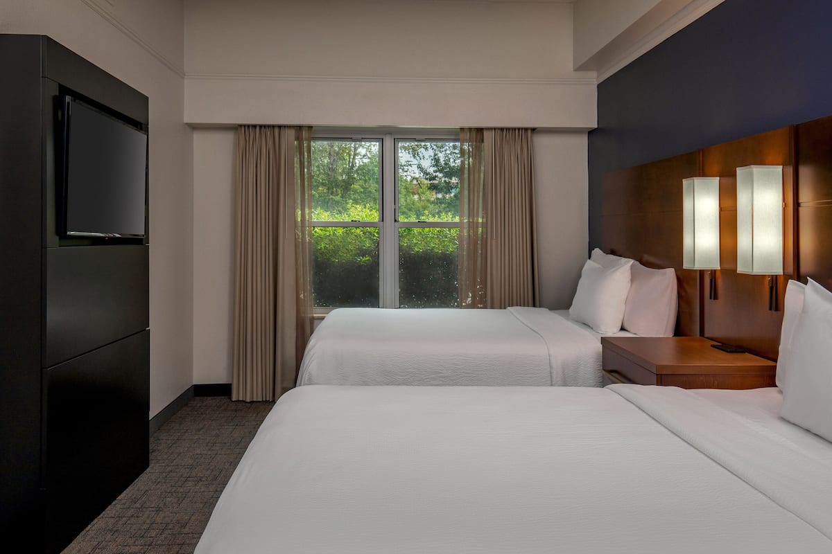 A hotel room with two beds, a TV, a nightstand with lamps, and a large window with curtains.