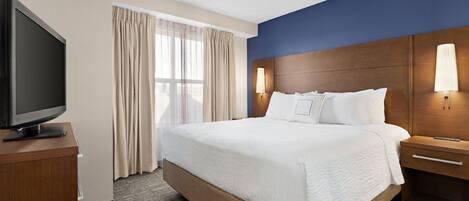 A hotel room with a large bed, a television on a wooden stand, a nightstand with a lamp, and a window with curtains.