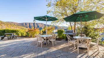 An outdoor patio with a stone floor, picnic tables, and chairs, surrounded by trees and a view of mountains.