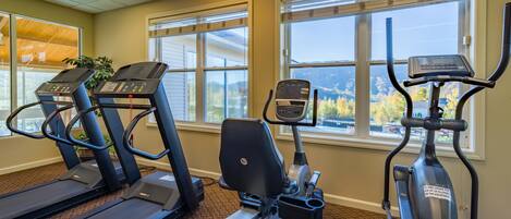 Ein Fitnessstudio mit Laufbändern, Crosstrainern und einem Ergometer, mit großen Fenstern und Blick ins Freie.