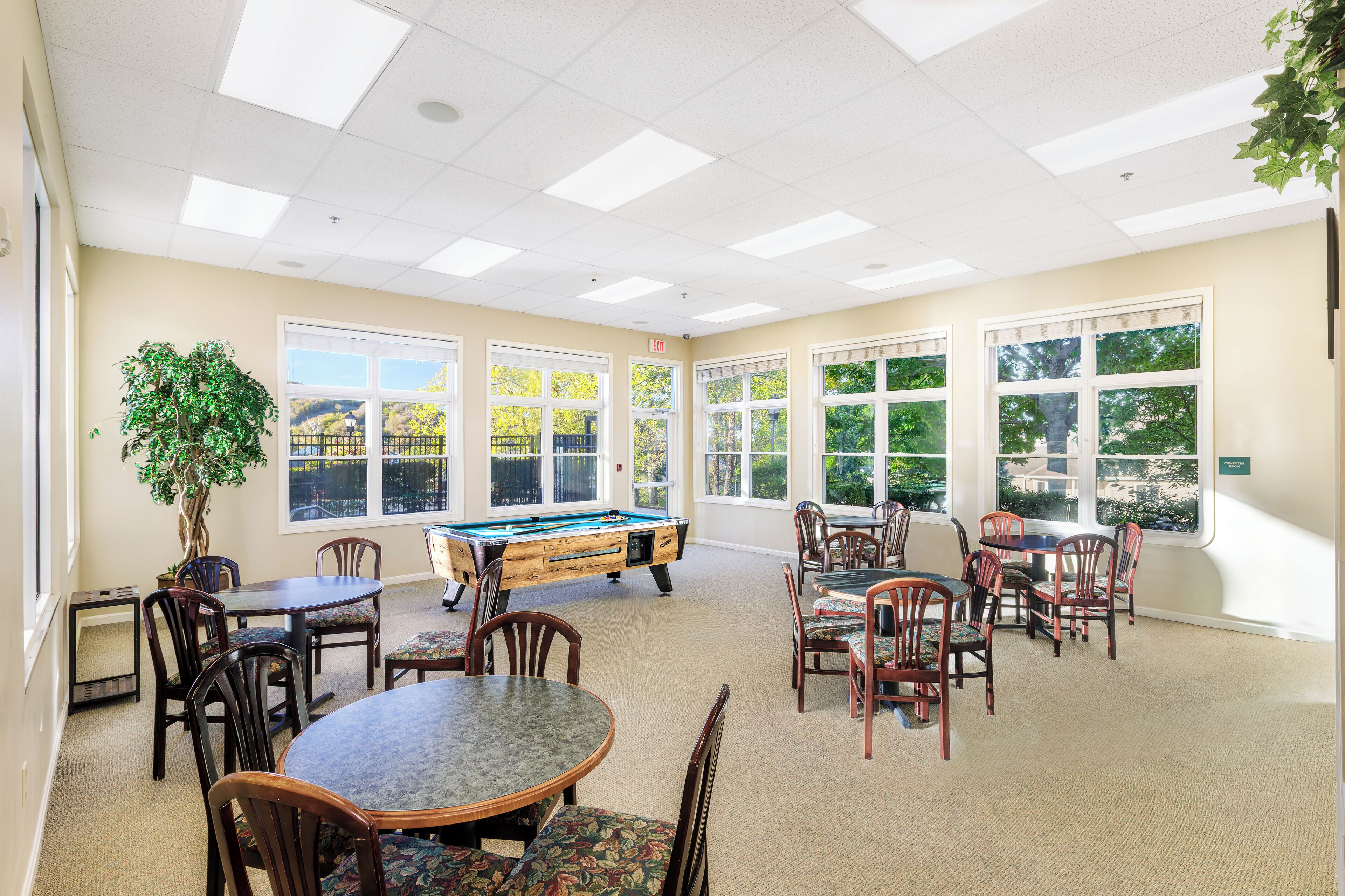 A room with a pool table, multiple dining tables, chairs, and large windows with a view of trees.