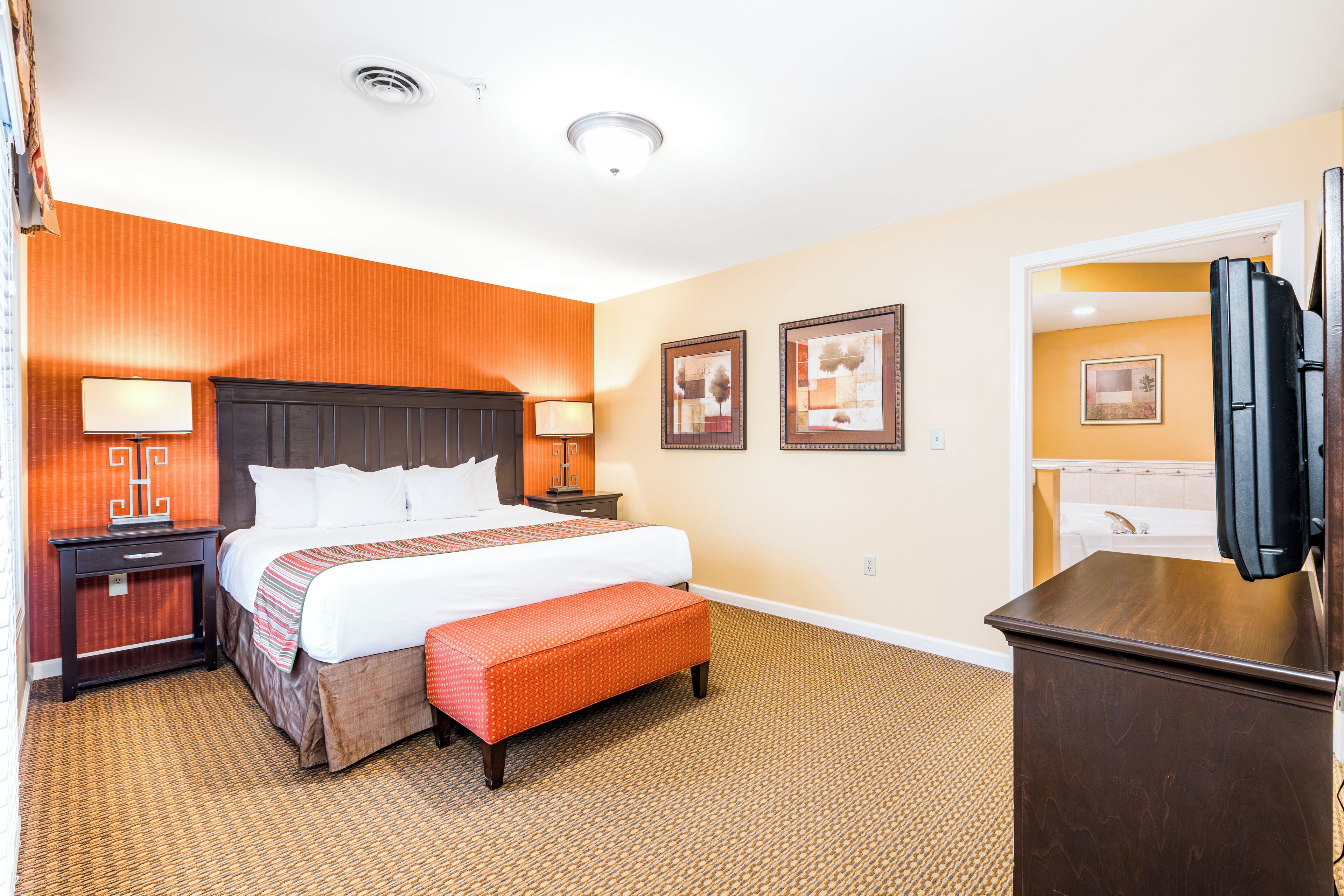 A hotel room with a large bed, a nightstand, a bench, a TV, and framed pictures on the wall.