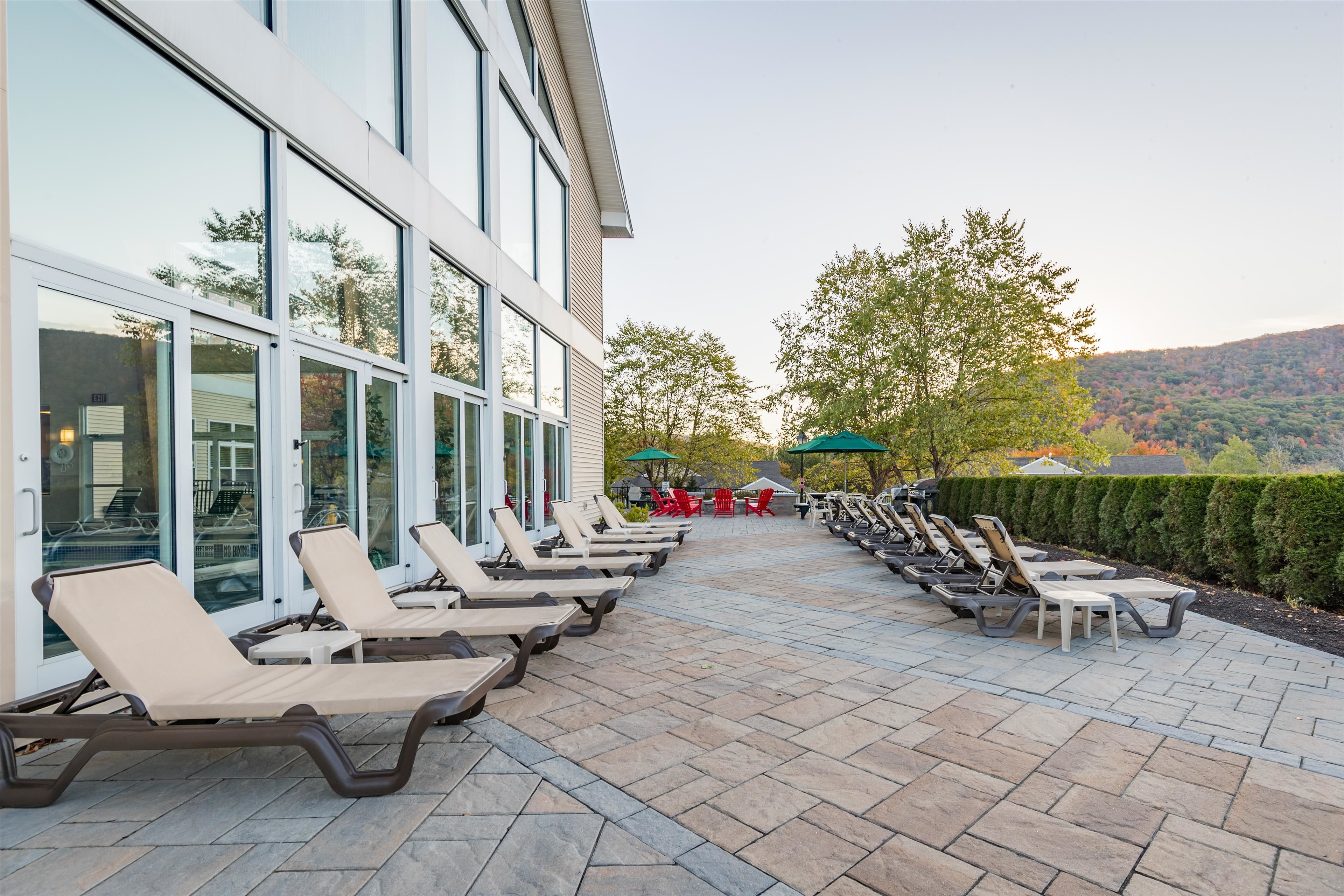A poolside area with lounge chairs and umbrellas, a building with large windows, and a view of trees and hills.