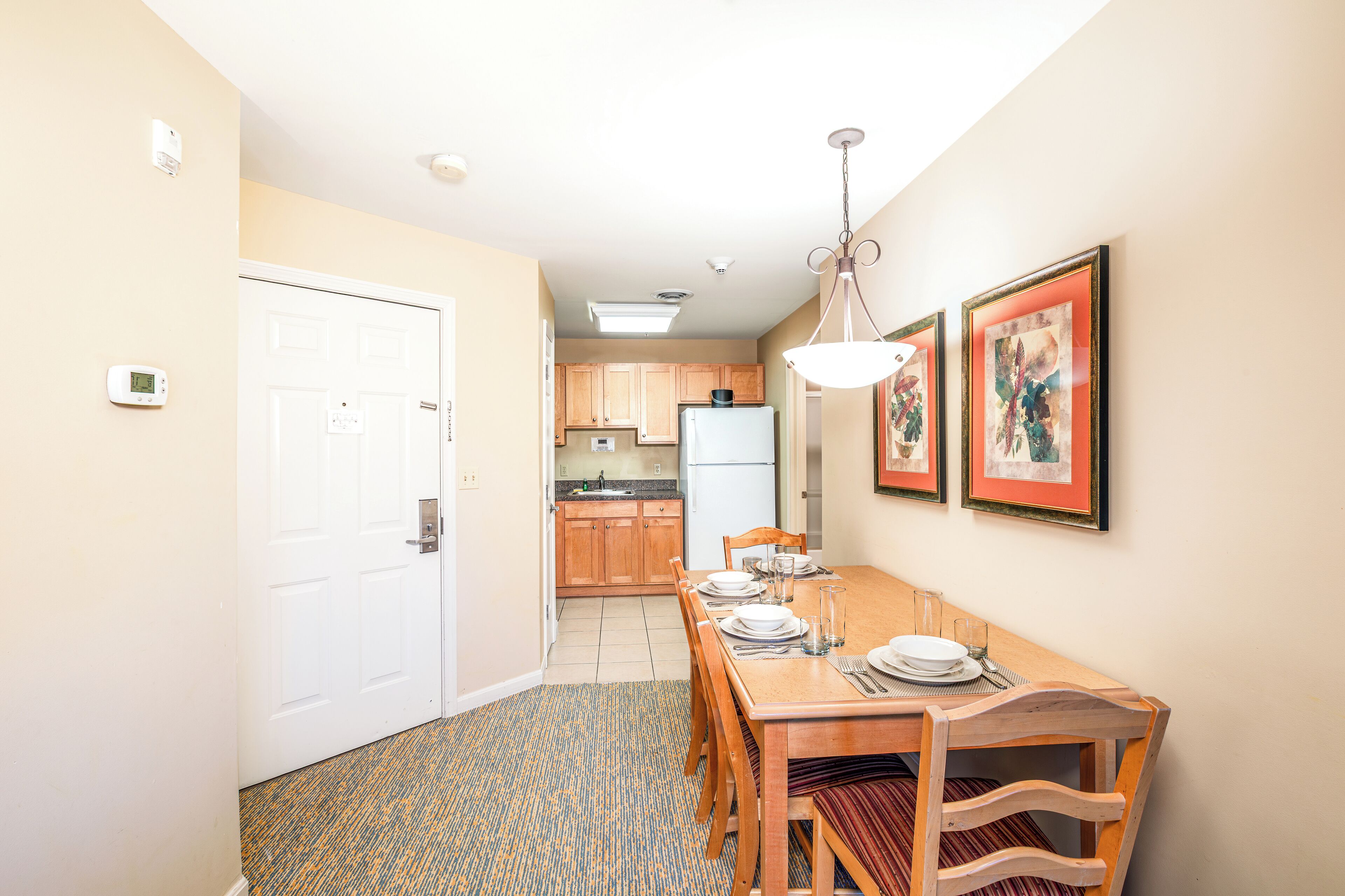 A dining area with a wooden table set for four, a kitchen in the background, and a door leading to another room.