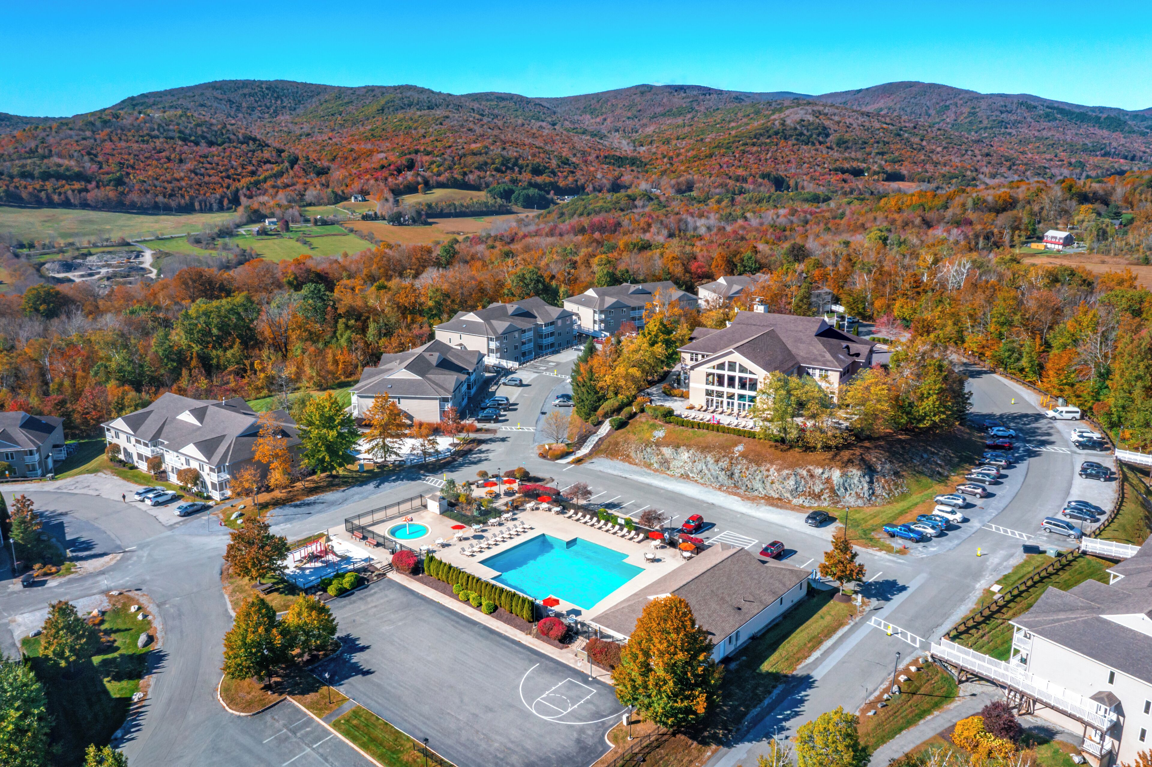 An aerial view of a residential area with a swimming pool, parking, and houses surrounded by autumn foliage.