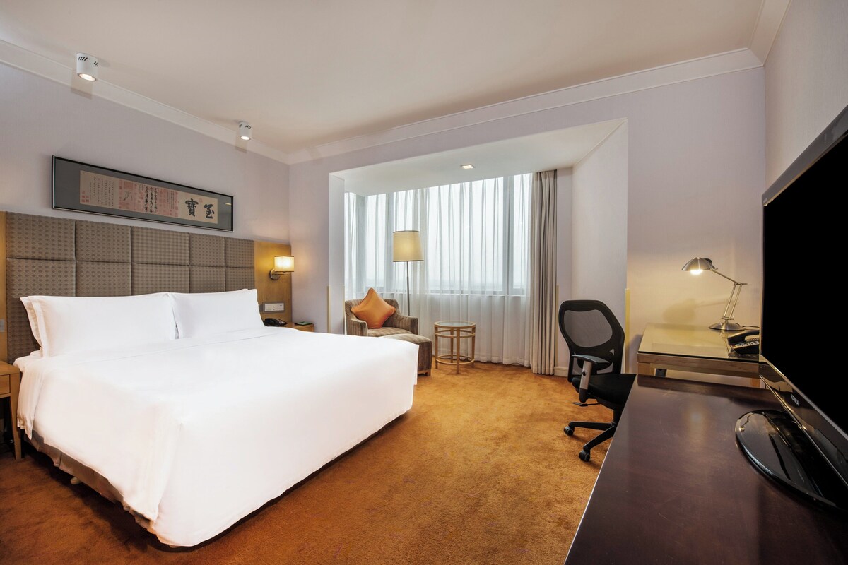 A hotel room with a large bed, a desk, a chair, a TV, and a window with curtains.