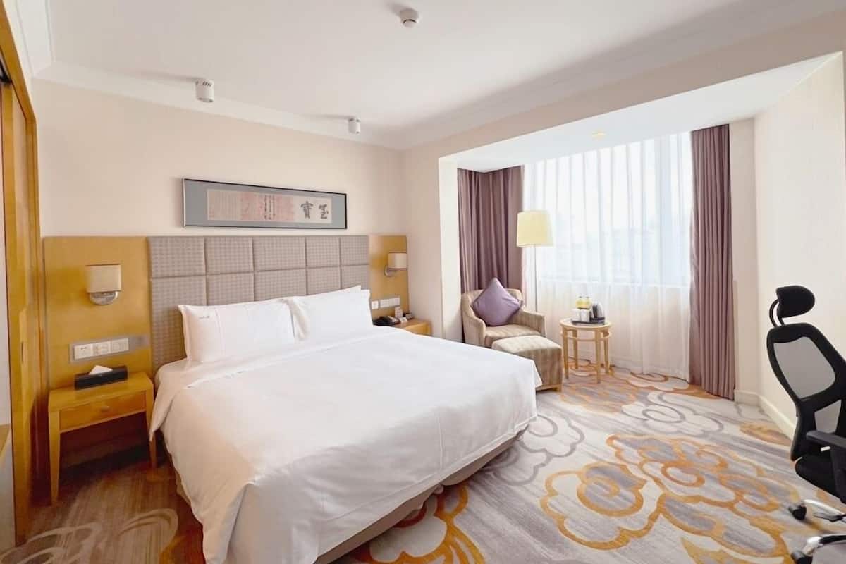 A hotel room with a large bed, a desk, a chair, a lamp, and a window with curtains.
