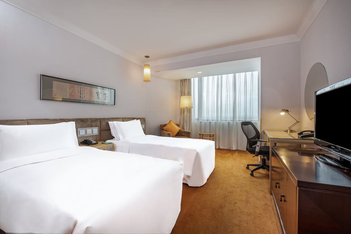 A hotel room with two beds, a desk, a TV, and a window with curtains.