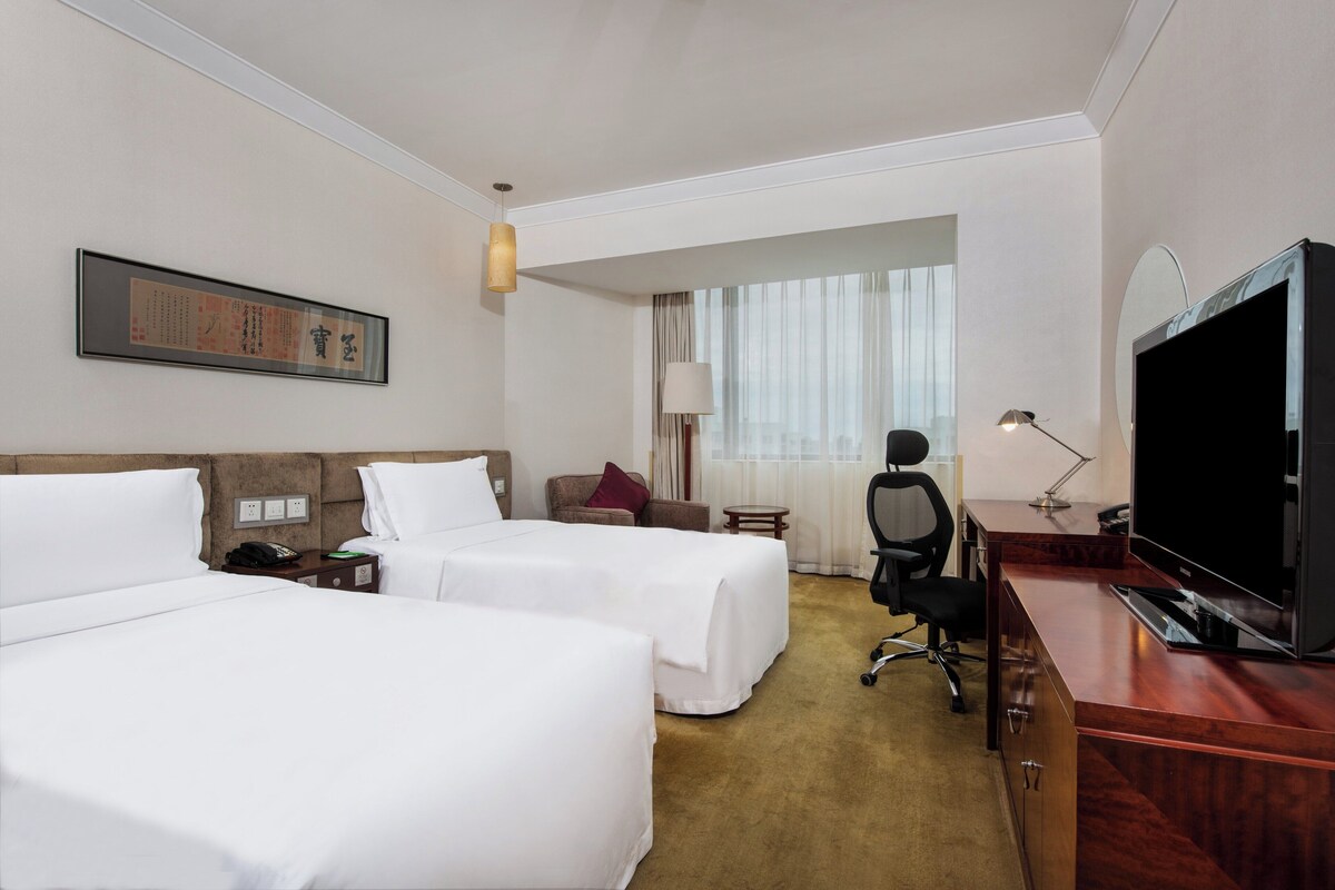 A hotel room with two beds, a desk with a TV, a chair, a lamp, and a window with curtains.