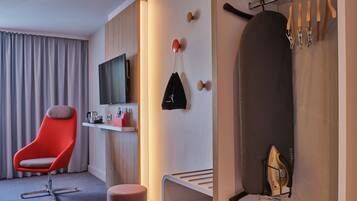 Moderni hotellihuone, jossa on taulutelevisio, punainen tuoli, pieni pöytä, jonka päällä on punainen esine, sekä silityslauta ja henkarit.