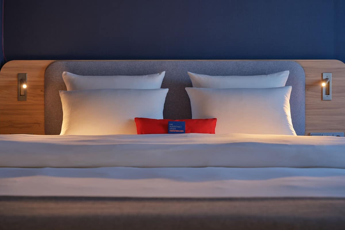 A neatly made bed with white linens and two pillows, a red accent pillow, and a blue tag in the center.