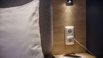 Una cama bien hecha con un almohadón blanco, una mesita de noche con un teléfono inteligente y su cargador, y un enchufe de pared.