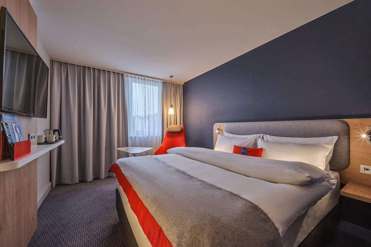 A hotel room with a large bed, a TV mounted on the wall, a desk with a chair, and a window with curtains.