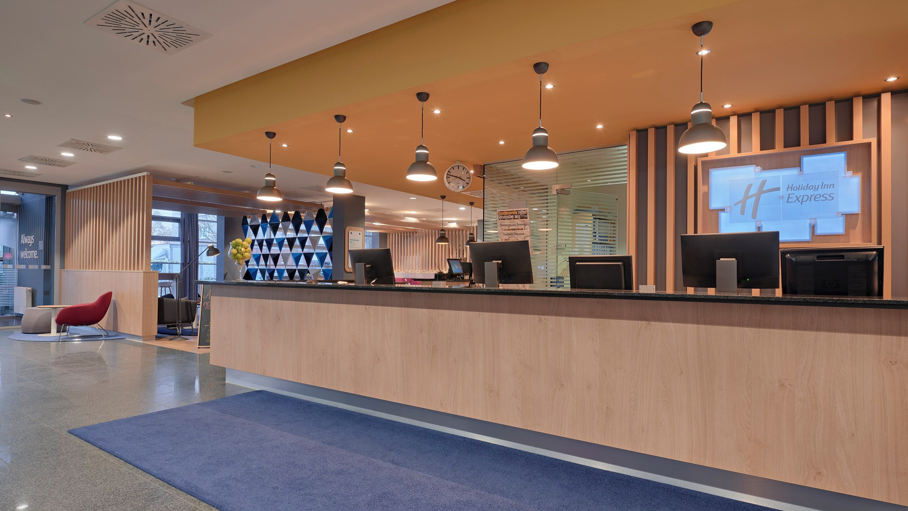 Un comptoir d’accueil d’hôtel au design moderne, comprenant un comptoir en bois, plusieurs écrans d’ordinateur et des luminaires suspendus.