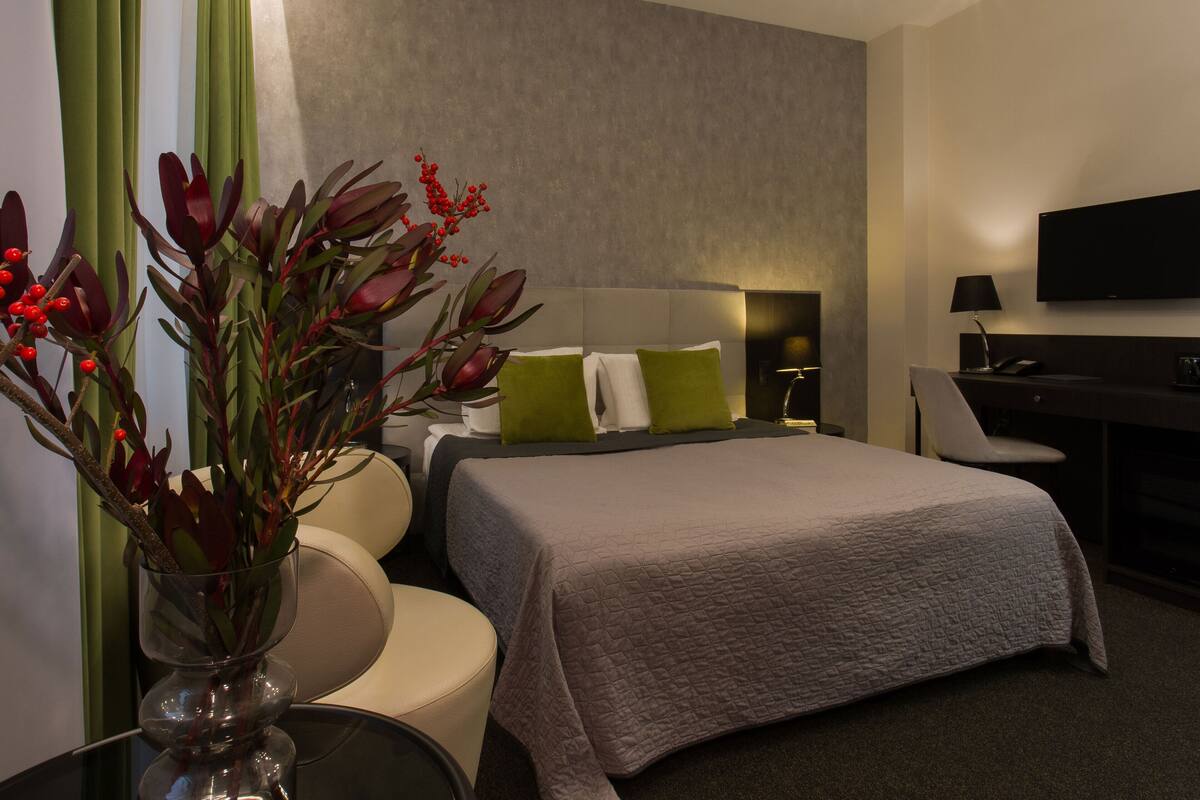 A modern hotel room with a large bed, a desk, a chair, a TV, and a vase with red flowers.