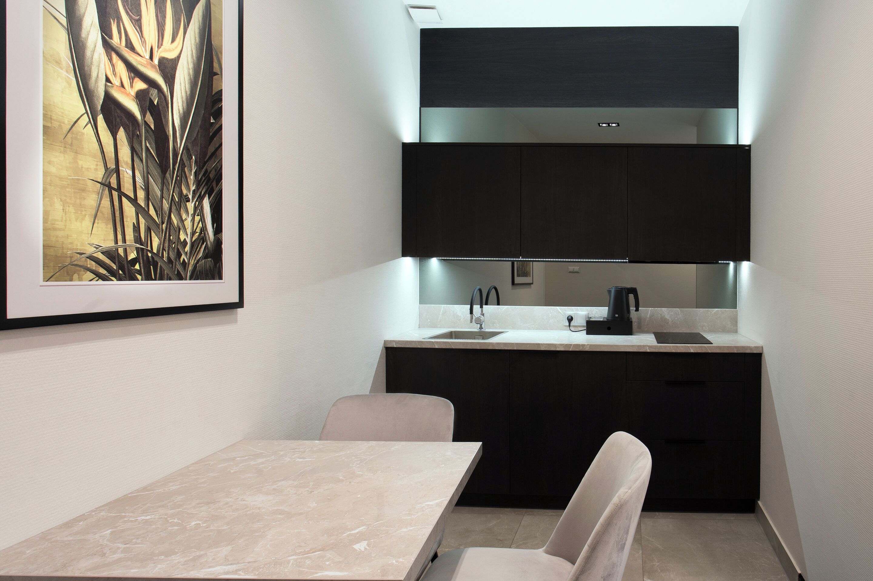 Una cocina moderna con armarios oscuros, encimera de mármol y un lavamanos. Se observa una obra de arte enmarcada sobre el lavamanos y una silla frente a la encimera.