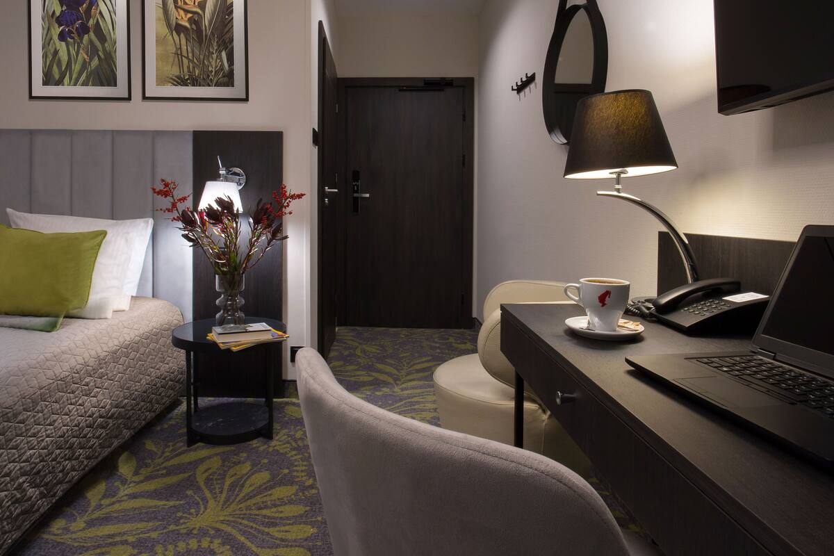 A hotel room with a bed, a desk with a laptop, a chair, a lamp, a cup of coffee, and two framed pictures on the wall.