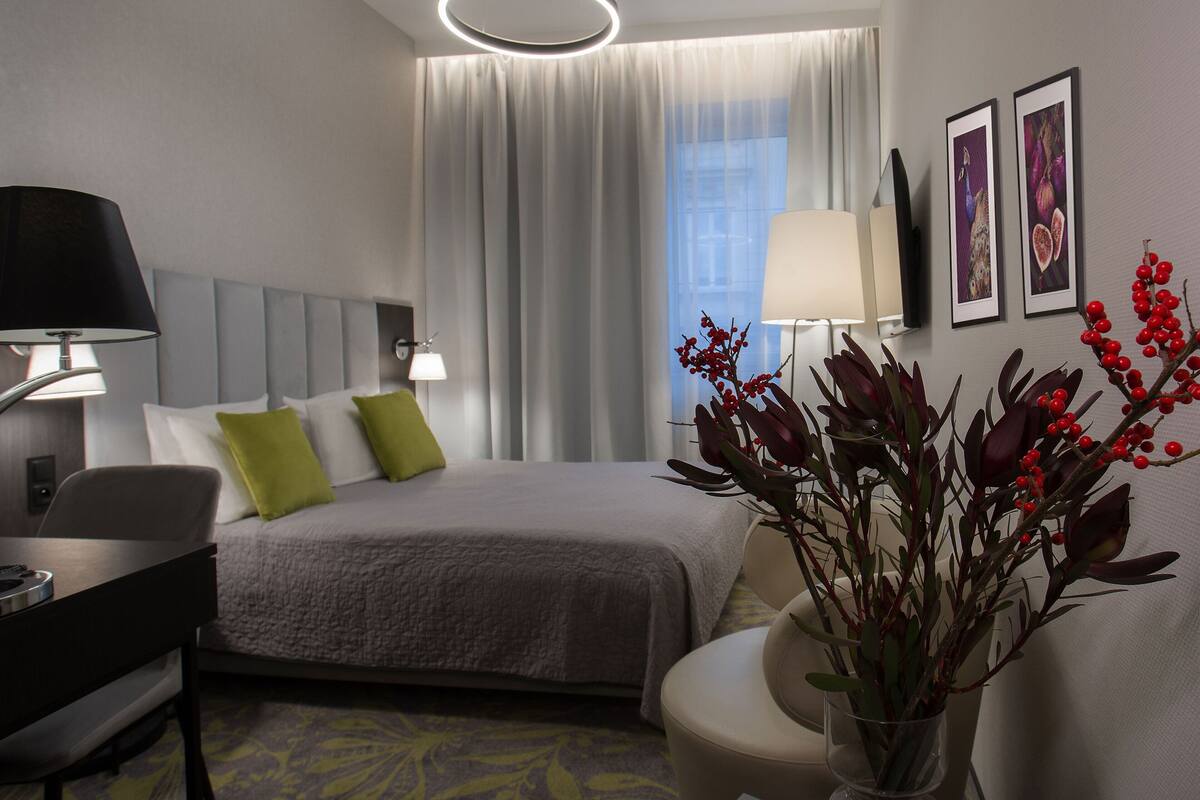 A hotel room with a bed, bedside table, lamp, chair, and a vase with flowers.