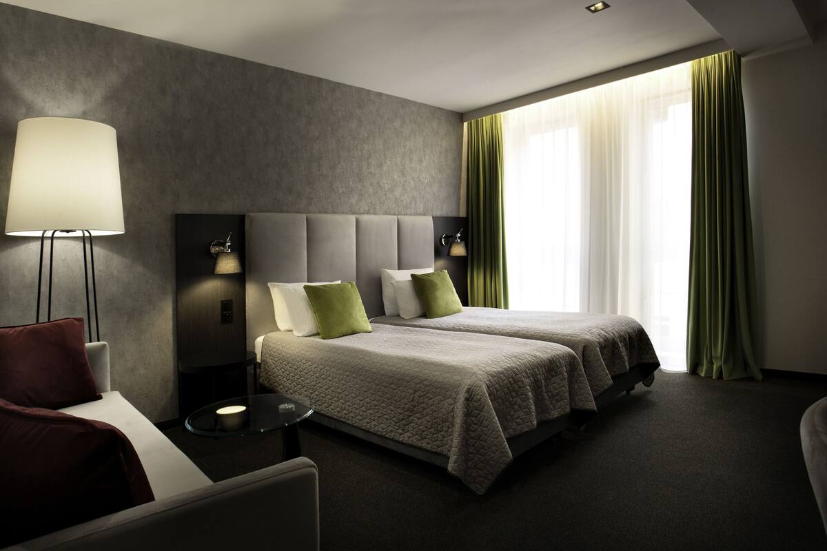 A hotel room with two beds, a desk, a chair, a lamp, and a window with curtains.