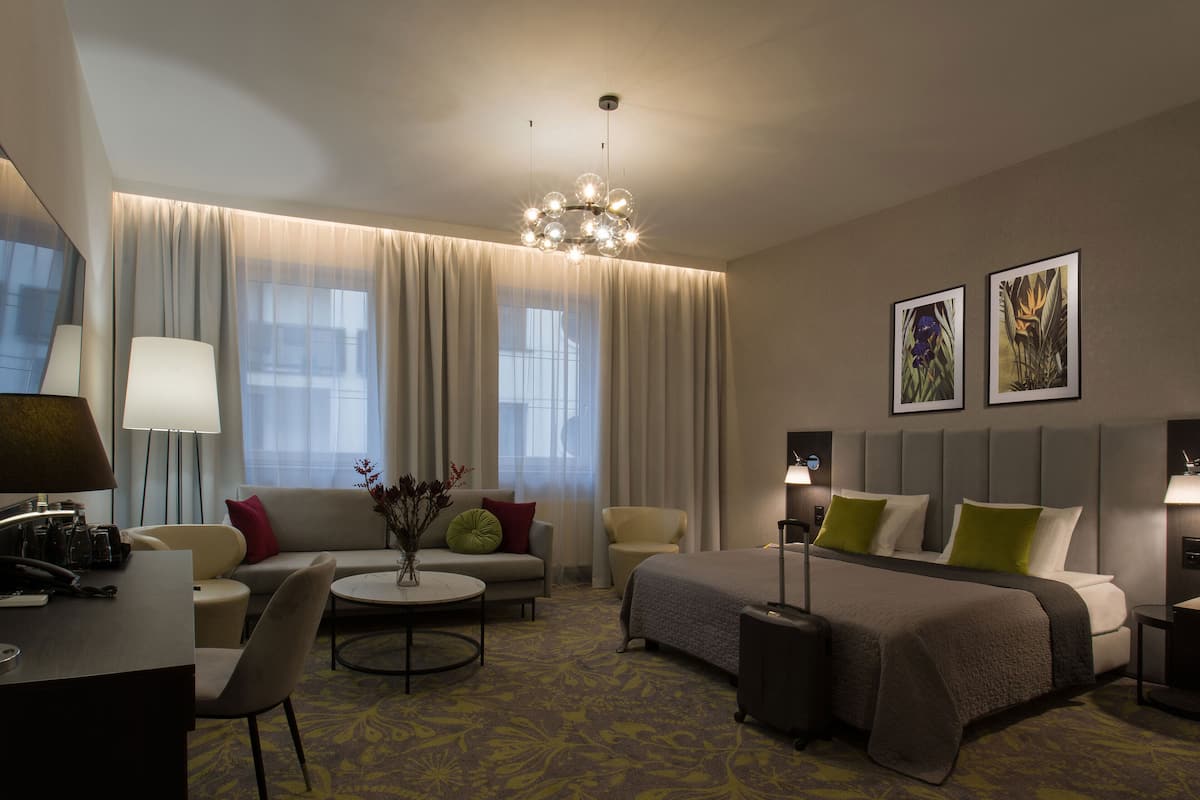 A hotel room with a bed, a sofa, a desk, a chair, a lamp, and a vase with flowers.
