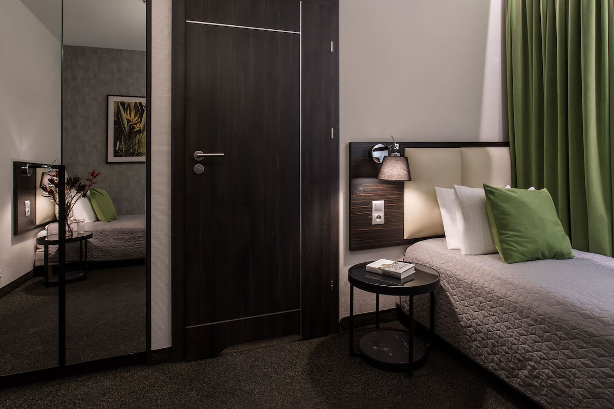 A modern hotel room with a dark wood wardrobe, a bedside table with a lamp, a green pillow, and a bed with a quilted cover.
