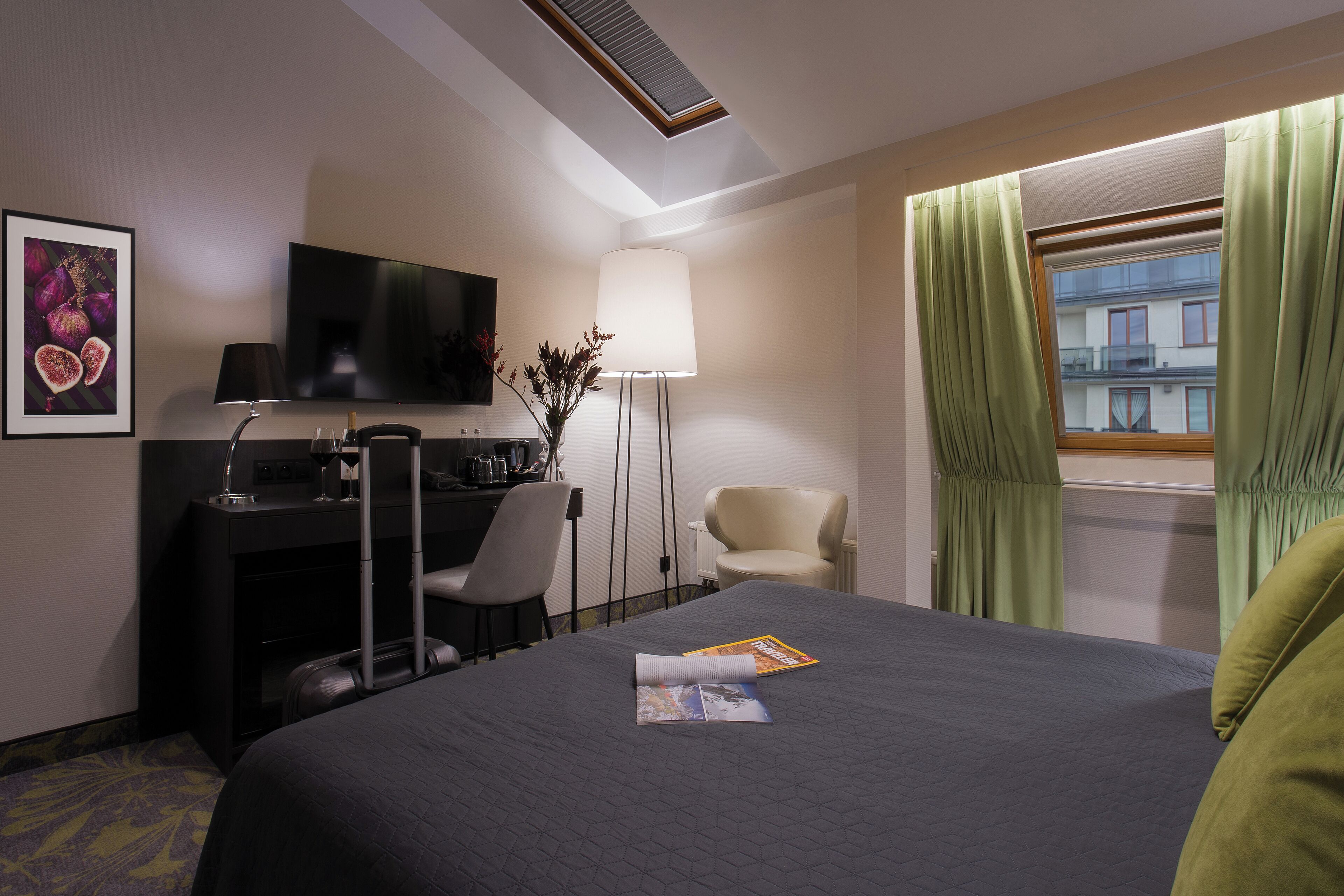 A hotel room with a bed, a desk with a computer, a chair, a TV, and a window with curtains.