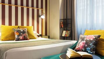 Une chambre d’hôtel avec un mur à rayures, un lit agrémenté d’oreillers jaunes et à motifs floraux, une table de chevet avec un livre, et une petite table d’appoint avec un livre.
