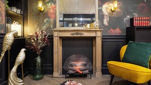 Et værelse med pejs, en gul lænestol, en grøn pude, en dekorativ fugleskulptur, en vase med blomster og en reol med bøger og pynteting.