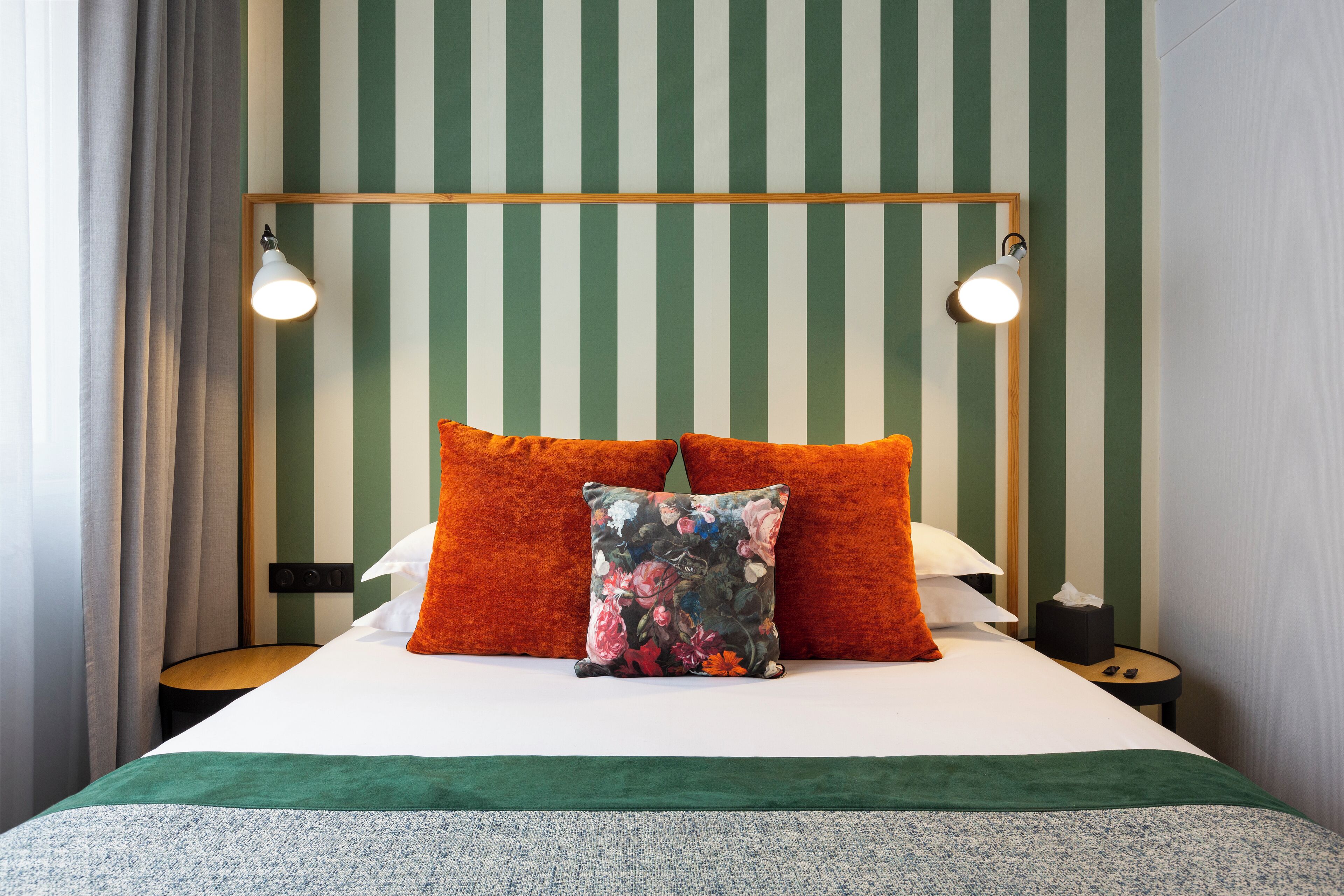 A neatly made bed with two pillows, a floral throw pillow, and a green bedspread. There are two bedside tables with lamps and a small nightstand with a box.
