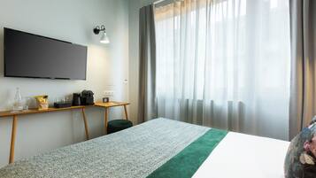 A hotel room with a bed, a flat-screen TV mounted on the wall, a small desk with a lamp, and a window with curtains.