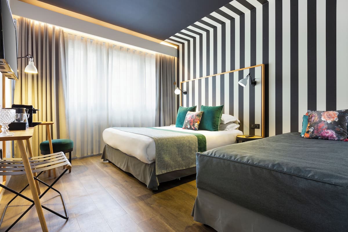Une chambre d’hôtel avec deux lits, un bureau et une chaise. Il y a une grande fenêtre avec des rideaux, un mur à rayures et une lampe sur le bureau.