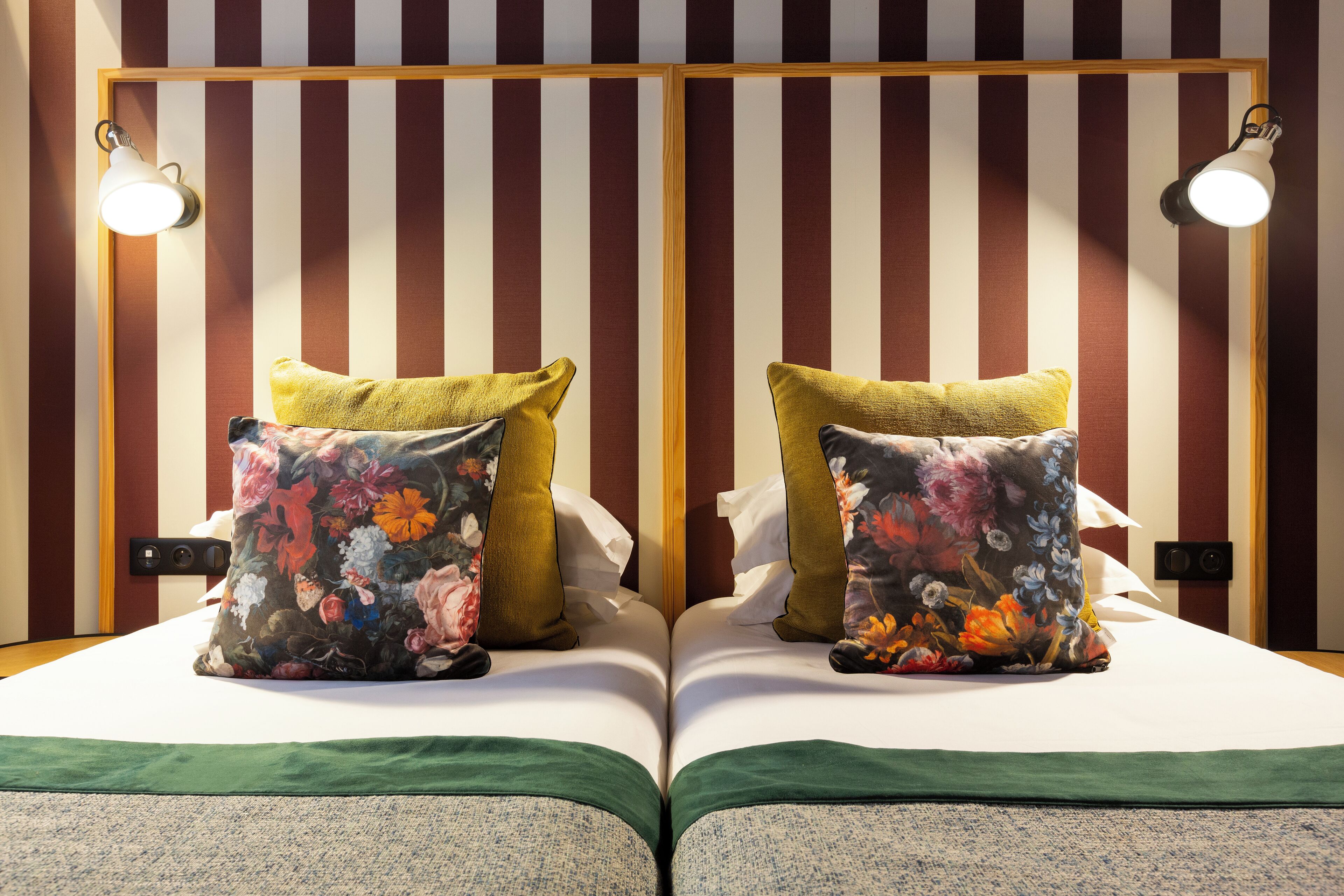 A double bed with floral pillows, a green bedspread, and a striped wall with two wall-mounted lights.