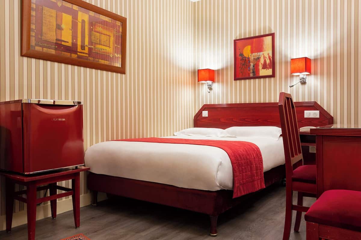 A hotel room with a bed, a desk, a chair, a TV, and two wall-mounted lamps.