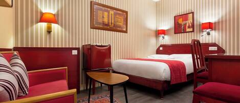 Una habitación de hotel con una cama, un sofá rojo, una mesita y dos lámparas de pared.